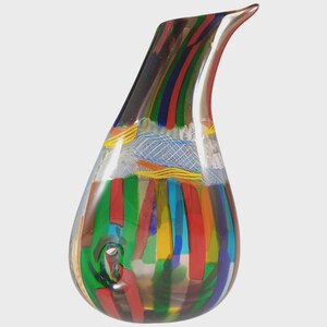 May include: A decorative art glass pitcher with a curved spout. The pitcher is adorned with vertical stripes in red, green, yellow, and blue, and has a clear glass handle. The design incorporates opaque and transparent sections, creating a vibrant decorative piece.