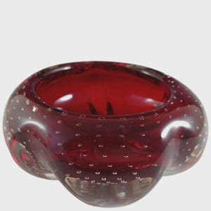 May include: A deep red glass bowl featuring a rounded, organic shape with a thick rim. The glass is embedded with numerous small, evenly distributed air bubbles throughout its structure, creating a textured appearance within the smooth surface.