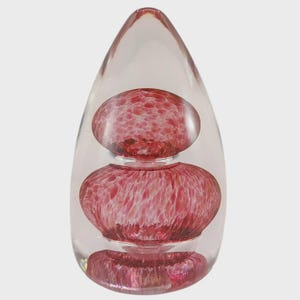 May include: A clear glass paperweight with a teardrop shape. Inside, three rounded, textured, red-pink glass forms are stacked. The top and bottom forms are smaller than the middle one. The paperweight is a decorative art object.