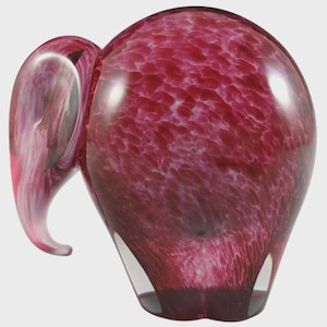 May include: A decorative glass elephant sculpture in shades of burgundy and white. The elephant has a rounded body and a curved trunk. The glass has a speckled pattern, and the sculpture is set against a neutral background.