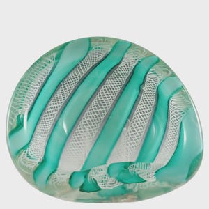 May include: A clear, oval glass paperweight featuring a turquoise and white striped design. Thick, diagonal turquoise stripes alternate with white, net-like patterns. The glass has a smooth, polished appearance, with a rounded shape.