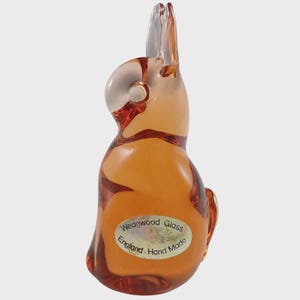 May include: A clear, amber-colored glass rabbit figurine. The rabbit has long ears and a rounded body. A sticker on the base reads "Wedgwood Glass England Hand Made."