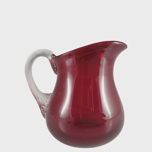 May include: A deep red glass jug with a clear glass handle. The jug has a rounded body and a pouring spout. The handle is curved and attached to the side. Likely used for serving drinks, the jug is a classic design.