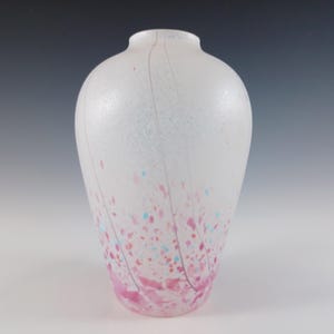 May include: A white glass vase with a speckled design. The vase features pink, blue, and red speckles, with pink streaks running down the sides. The vase has a rounded body and a narrow neck, suitable for floral arrangements or decorative display.