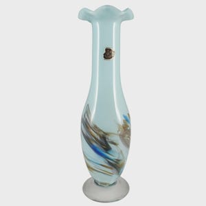May include: A tall, pale blue glass vase with a frilled top and a clear, circular base. The vase has swirling patterns in brown, blue, and white. A small, gold-toned label is attached. This decorative vase is suitable for floral arrangements.