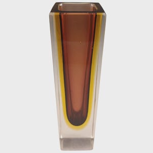 May include: A tall, square-shaped glass vase featuring a layered design with a clear outer layer, a vibrant yellow middle section, and a deep brown inner core. The vessel has a smooth, polished finish and a tapered silhouette that narrows towards the base.