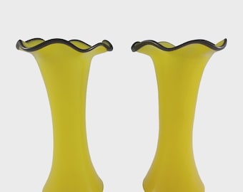 May include: Two yellow glass vases with a ruffled rim and a dark brown trim. The vases have a slender, elongated shape, widening slightly at the base. The vases are a bright, cheerful yellow color.