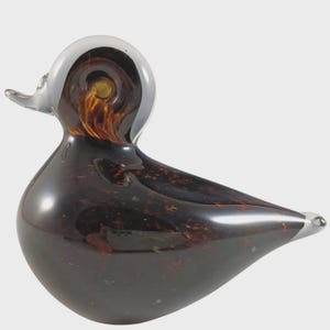 May include: A decorative glass duck figurine. The duck has a clear head and beak, with a dark brown body speckled with reddish-brown inclusions. The duck's head has a swirl pattern with a yellow center.