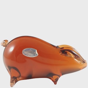 May include: A brown glass pig figurine with a glossy finish. The pig has small legs, a curled tail, and a pointed snout. A small, oval sticker on the side reads "Wedgwood England, Hand Made."