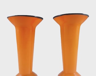 May include: Two orange vases with a flared top and a black rim. The vases have a cylindrical body and a rounded base. The background is a light gray.