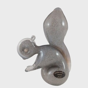 May include: A gray crystal squirrel figurine with a large, bushy tail. The squirrel is in a seated position, with its paws resting on a round base. The figurine has a speckled pattern and a clear glass head. The base has a black label that reads "Wedgwood Crystal Ltd Handmade England."