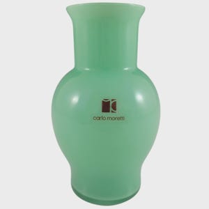 May include: A pale green glass vase with a bulbous base and a fluted neck. The vase has a smooth finish and a brown logo that reads "carlo moretti". A decorative item for the home.