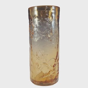 May include: A tall, textured glass vase with a golden-amber gradient. The vase has a rough, organic texture and a slightly flared rim. The glass appears to be thick and sturdy, suitable for displaying flowers or as a decorative piece.