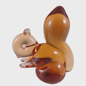 May include: A glass squirrel figurine in shades of amber and brown. The sculpture features a rounded body, a bushy tail, and small, detailed paws. The squirrel is posed in a seated position, with its head turned slightly to the side.