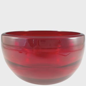 May include: A deep, translucent red glass bowl with a smooth, rounded silhouette and thick walls. The bowl features a wide, open top and a curved base, showcasing a vibrant crimson hue throughout the material with subtle light reflections.