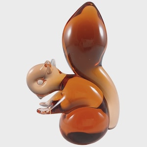 May include: A glass squirrel figurine in shades of brown and tan. The squirrel is posed with its head down and tail up. The figurine has a smooth, glossy finish and is likely a decorative item.