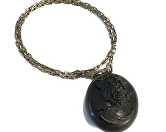 May include: A round black pendant featuring a raised profile portrait of a person with curly hair, attached to a metallic chain. The pendant has a smooth, dark finish and the portrait is detailed with texture, suspended from a brass-toned link chain.