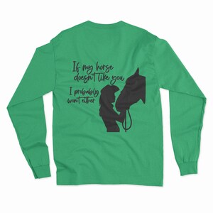 May include: Green long-sleeved t-shirt with a black silhouette of a woman and a horse. The text on the shirt reads "If my horse doesn't like you I probably won't either."