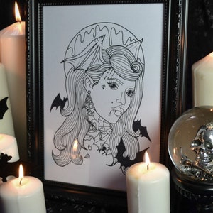 Peut inclure: Une impression encadrée noire d'un dessin au trait d'une femme avec des ailes de chauve-souris et un motif de toile d'araignée. L'impression est entourée de bougies blanches et d'un globe à neige en verre avec un crâne argenté à l'intérieur.