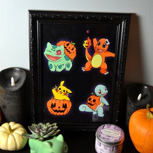 May include: A black framed print with a white background featuring five cartoon characters from the Pokemon franchise dressed in Halloween costumes. The characters are Bulbasaur, Charmander, Pikachu, Squirtle, and a pumpkin.