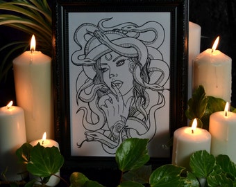 May include: Black and white line art print of a woman with snakes for hair. The woman is looking down and has her finger to her lips. The print is framed in a black frame and is surrounded by candles and greenery.