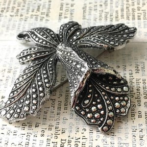 May include: A silver-coloured metal brooch shaped like a flower. The brooch has detailed patterns with small, round decorations on the petals and leaves. The pin is set against a vintage paper background.