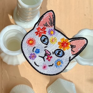 May include: White embroidered iron-on patch of a cat's face with colourful flowers on its fur.