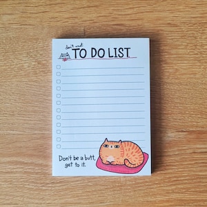 Puede incluir: Un bloc de notas blanco con una línea roja y el texto "TO DO LIST" escrito en negro. El bloc de notas tiene una lista de verificación con 10 casillas vacías. La parte inferior del bloc de notas tiene un gato naranja de dibujos animados con rayas negras acostado sobre un cojín rosa. El texto "Don't be a butt, get to it." está escrito debajo del gato.