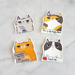 Peut inclure: Quatre autocollants de chats avec des expressions et des textes différents. Le premier autocollant montre un chat gris avec le texte "First, Coffee." Le deuxième autocollant montre un chat calico avec le texte "This is my happy face." Le troisième autocollant montre un chat orange avec le texte "Computer says no." Le quatrième autocollant montre un chat noir et blanc avec le texte "Feed me."