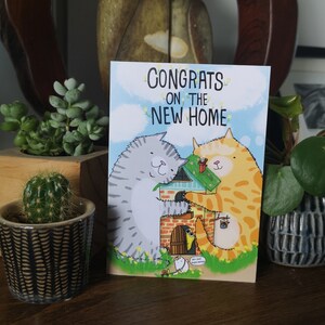 May include: A greeting card with a cartoon illustration of two cats building a house. The card reads "Congrats on the new home".