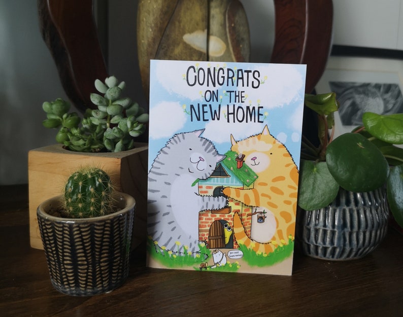 May include: A greeting card with a cartoon illustration of two cats building a house. The card reads "Congrats on the new home".