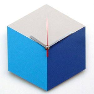 Pu&ograve; includere: Un orologio moderno con un design geometrico. Il quadrante dell'orologio &egrave; un esagono con tre lati in grigio chiaro, tre lati in blu e una lancetta delle ore rossa e una lancetta dei minuti argentata.