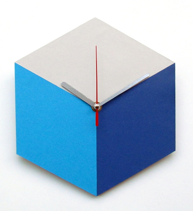Pu&ograve; includere: Un orologio moderno con un design geometrico. Il quadrante dell'orologio &egrave; un esagono con tre lati in grigio chiaro, tre lati in blu e una lancetta delle ore rossa e una lancetta dei minuti argentata.