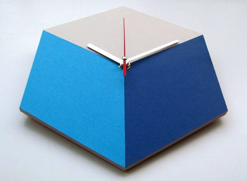 Pu&ograve; includere: Un orologio geometrico con un quadrante bianco, blu e blu chiaro. Le lancette dell'orologio sono argentate e rosse.