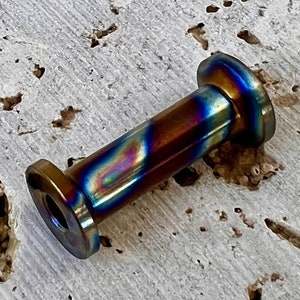 Può includere: Primo piano di un piccolo oggetto cilindrico in metallo iridescente con estremità arrotondate. L'oggetto presenta una combinazione di colori blu, viola e oro.