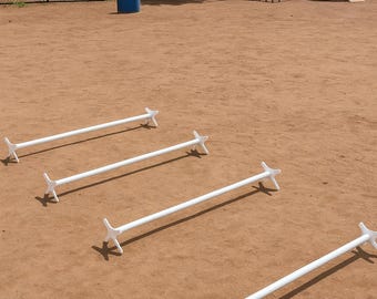 May include: An outdoor dog agility course with white jump bars. The bars are set on a dirt surface, with other agility equipment visible in the background, including weave poles and a ramp.