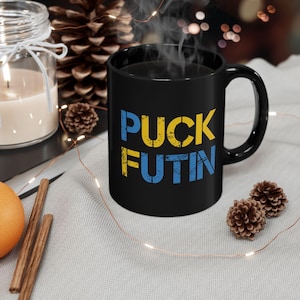 Może przedstawiać: Czarny kubek do kawy z napisem "PUCK FUTIN" w żółtych i niebieskich literach. Kubek stoi na białym tle z łańcuchem świateł.