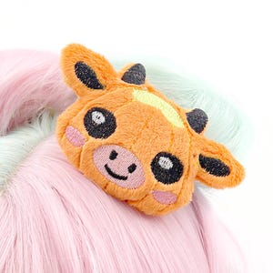 May include: An orange plush cow hair clip with black eyes, ears, and horns, and a smiling face. The cow clip is set against a backdrop of pink and light blue wig hair.