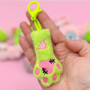 May include: A lime green plush cat paw keyring with pink paw pads and black stitches. The keyring has a neon green clip. Other plush keyrings are in the background on a pink surface.