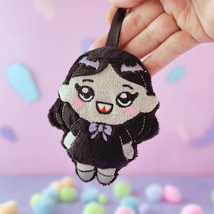 Pode incluir: Um porta-chaves de peluche com uma personagem estilizada de cabelo preto, olhos grandes e expressivos e um pequeno dente vermelho. A personagem usa um vestido preto com um laço roxo claro na gola e tem pele clara com bochechas rosadas.