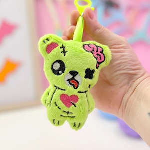 May include: A lime green plush bear keyring with zombie-themed details. The bear has a pink brain, a heart, and stitched details. It has a yellow clip for attaching to bags or keys. The background includes bat-shaped decorations.
