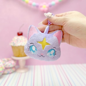 May include: A plush lavender cat-shaped keychain with embroidered blue eyes and a yellow star. The cat has pink inner ears and a small, black embroidered nose and whiskers. The keychain is held by a hand against a pink background with a floral garland.