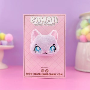 以下が含まれることがあります： 青い目をしたピンクと白のぬいぐるみ猫の頭のヘアクリップ。クリップは「Kawaii Hair Candy」というテキストとウェブサイトアドレスが書かれたピンクのカードの上にあります。