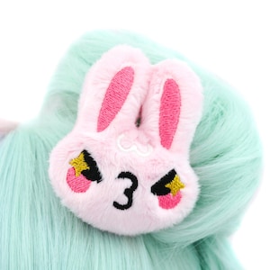Pu&ograve; includere: Un fermaglio per capelli in peluche rosa a forma di coniglio con occhi ricamati, il numero "3" e stelle gialle. Il coniglio ha un'espressione arrabbiata.