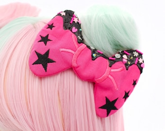 May include: A pink and black bow with a heart and star design. The bow is decorated with glitter and other embellishments.