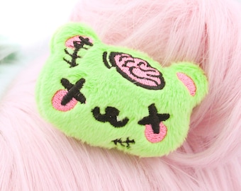 May include: Green plush bear with pink embroidered details, including a brain, eyes, and stitches. The bear is attached to a pink wig.