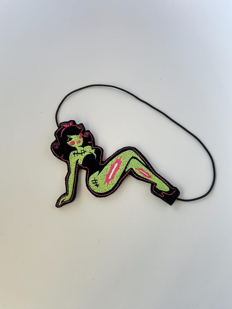 May include: An embroidered patch featuring a stylized green-skinned figure with black hair, wearing a black outfit. The figure has pink accents, visible stitches on the skin, and a pink wound detail on the leg. A thin black elastic cord is attached.