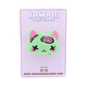 May include: Green plush cat hair clip with pink ears and a pink brain embroidered on the head. The clip has black eyes with an X over them. The clip is on a pink background with the text "Kawaii Hair Candy" and "www.kawaihaircandy.com".