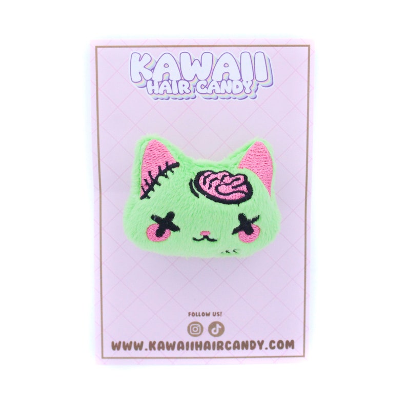 May include: Green plush cat hair clip with pink ears and a pink brain embroidered on the head. The clip has black eyes with an X over them. The clip is on a pink background with the text "Kawaii Hair Candy" and "www.kawaihaircandy.com".