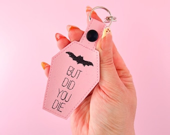 Puede incluir: Un llavero de color rosa claro con forma de ataúd que presenta un bordado negro de un murciélago y el texto BUT DID YOU DIE. El llavero está hecho de un material liso con costuras visibles y un cierre de botón a presión negro.